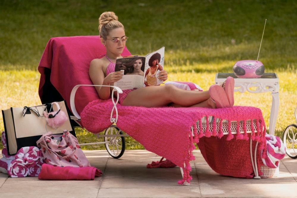 A woman in a pink bikini lies on a deck chair covered in pink blankets, reads a magazine. there are pink towels, a tote bag and a radio next to her.